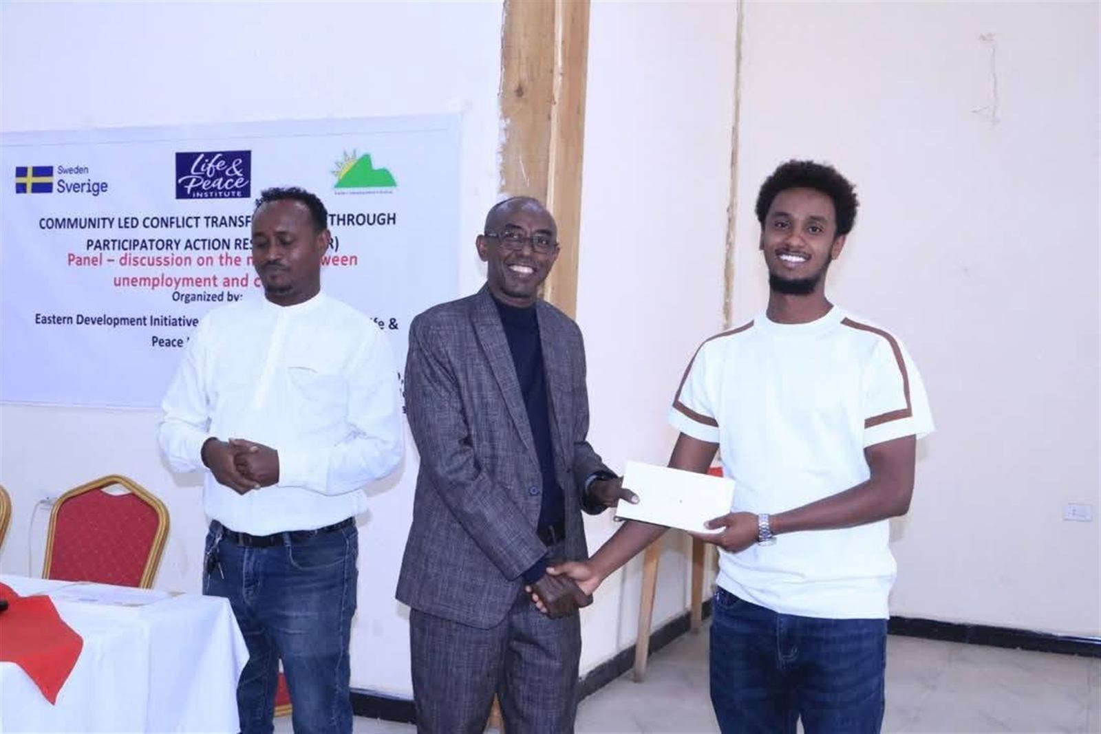 Three men shaking hands in front of the event banner, certificate handover moment