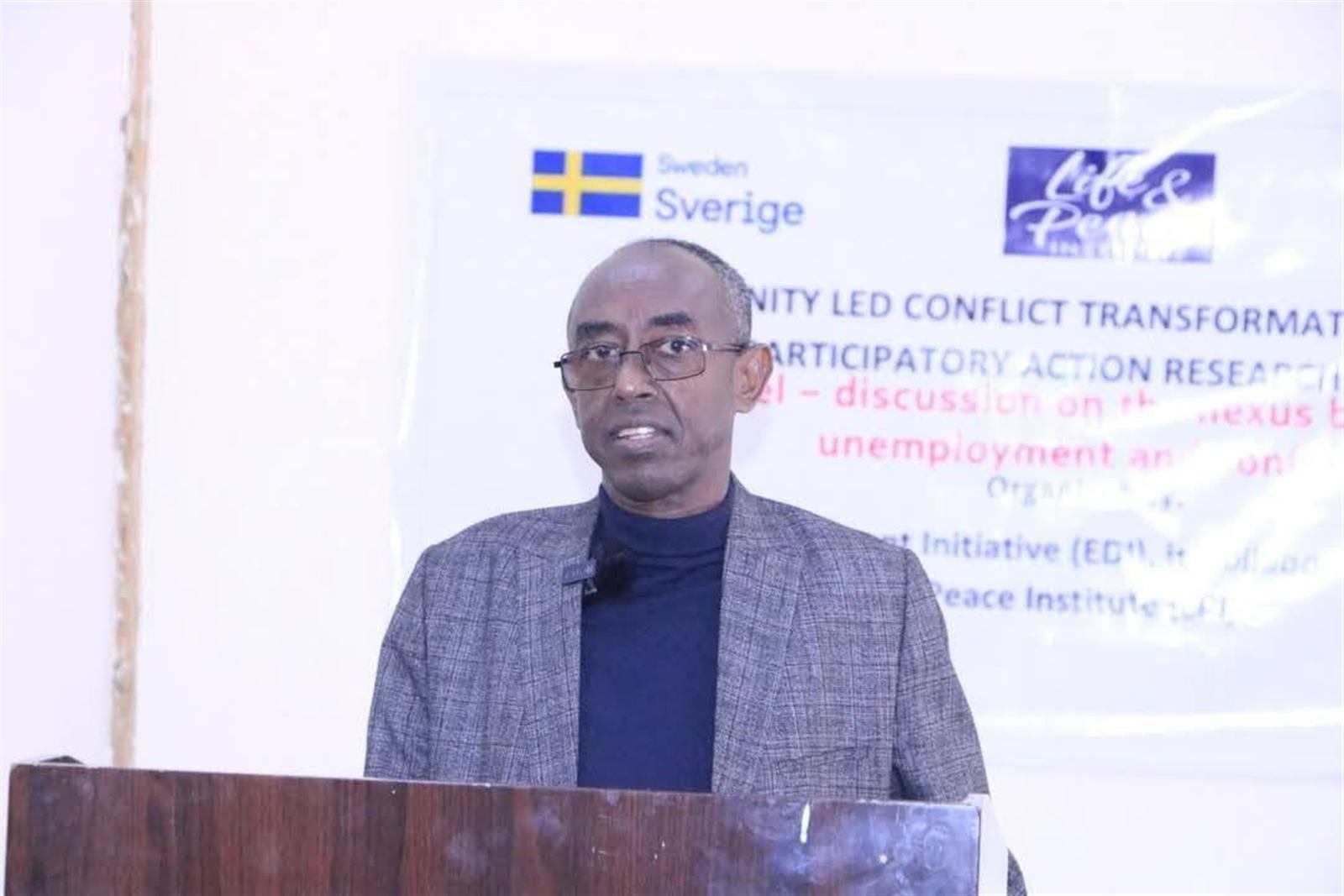 Speaker at the podium with banner text on unemployment and conflict visible behind him