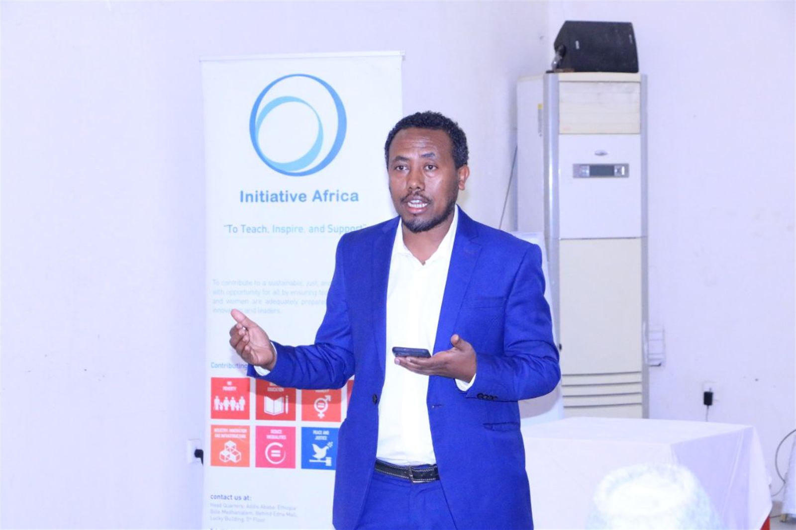 Speaker in a blue suit presenting beside an Initiative Africa partner-organization banner