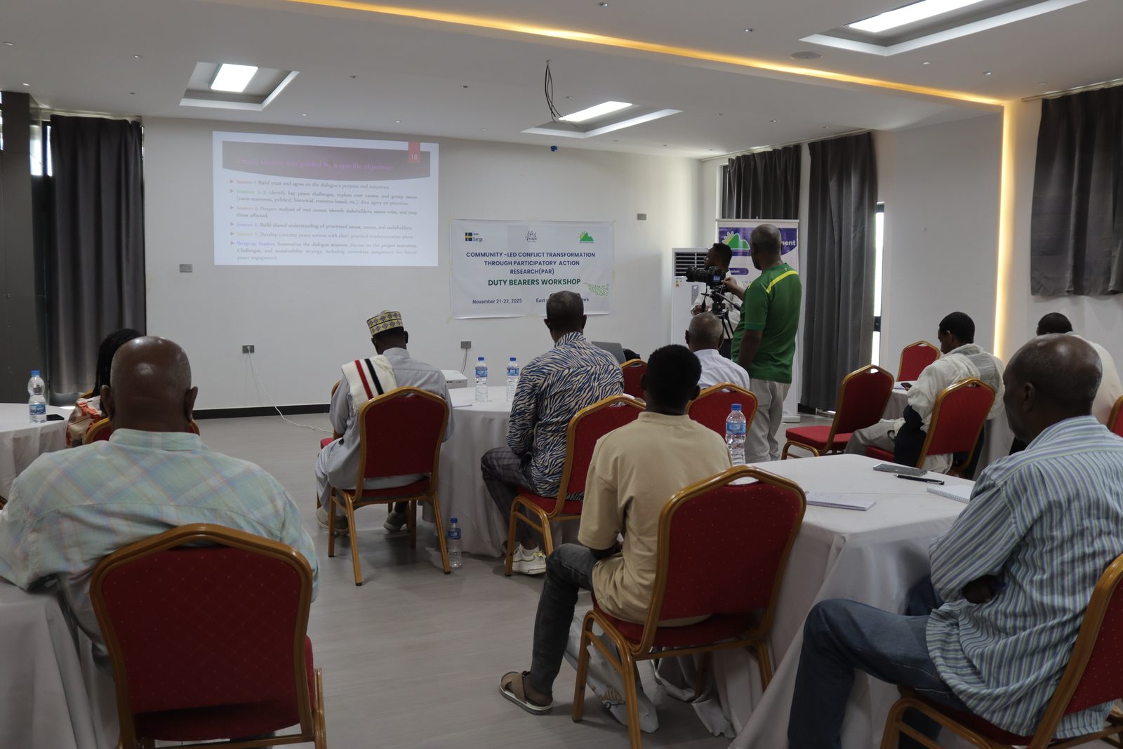 Wide room view: Duty Bearers Workshop banner and a slide on dialogue-session objectives, a video crew filming