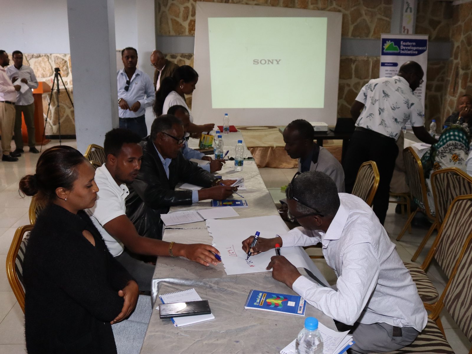 A participant writes on a flip chart during a working-group discussion, EDI rollup banner visible behind