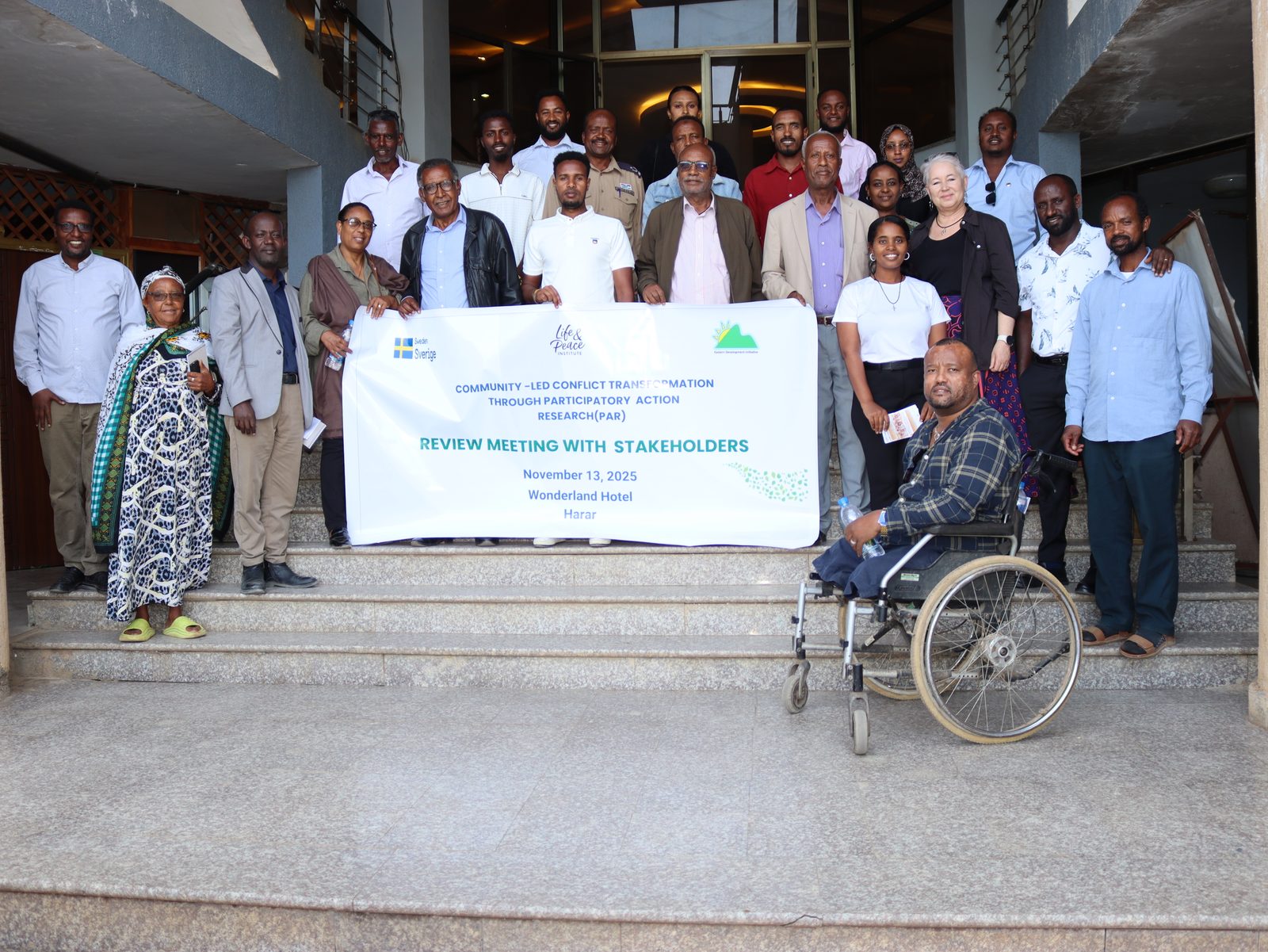 Full participant group photo on the hotel steps holding the event banner, a wheelchair user seated in front