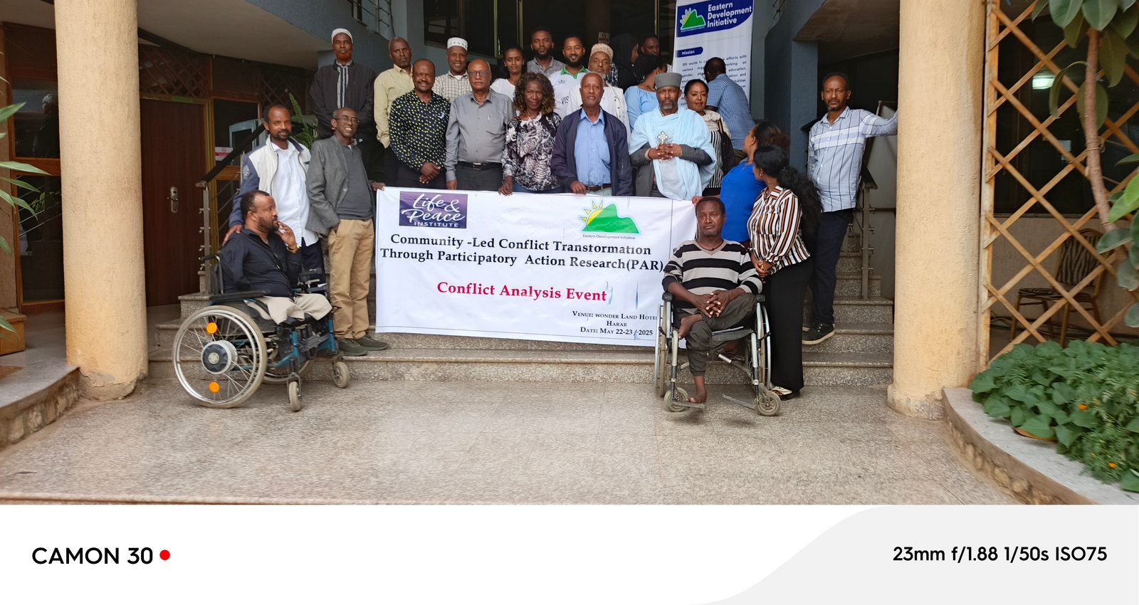 Outdoor group photo holding a banner with the full event name, venue, and date; two wheelchair users seated in front