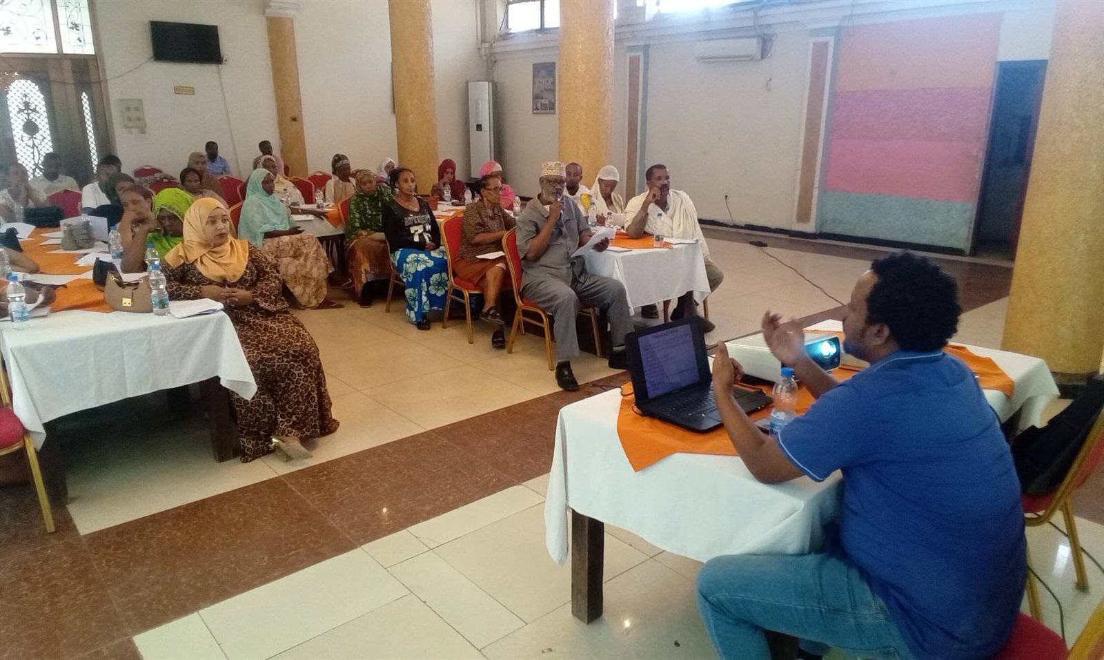 Facilitator at a laptop and projector addressing a seated circle of participants