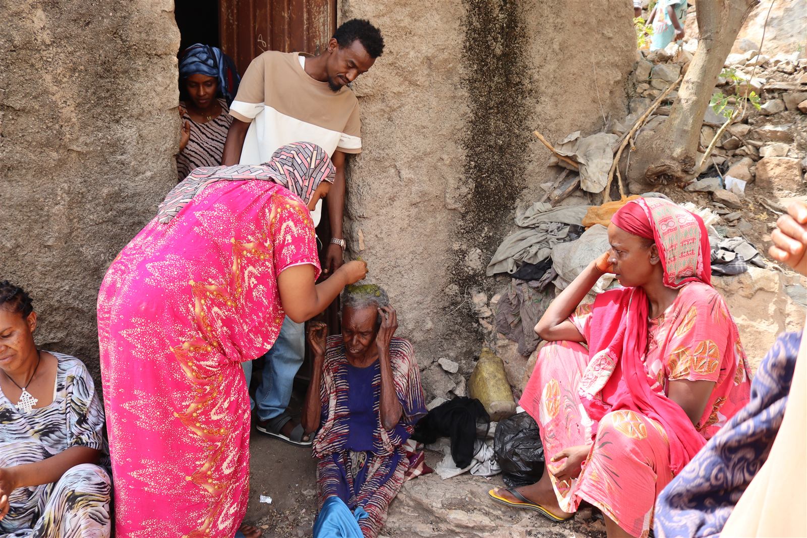 A woman in a pink dress washes the hair of a seated elderly woman at a stone doorway