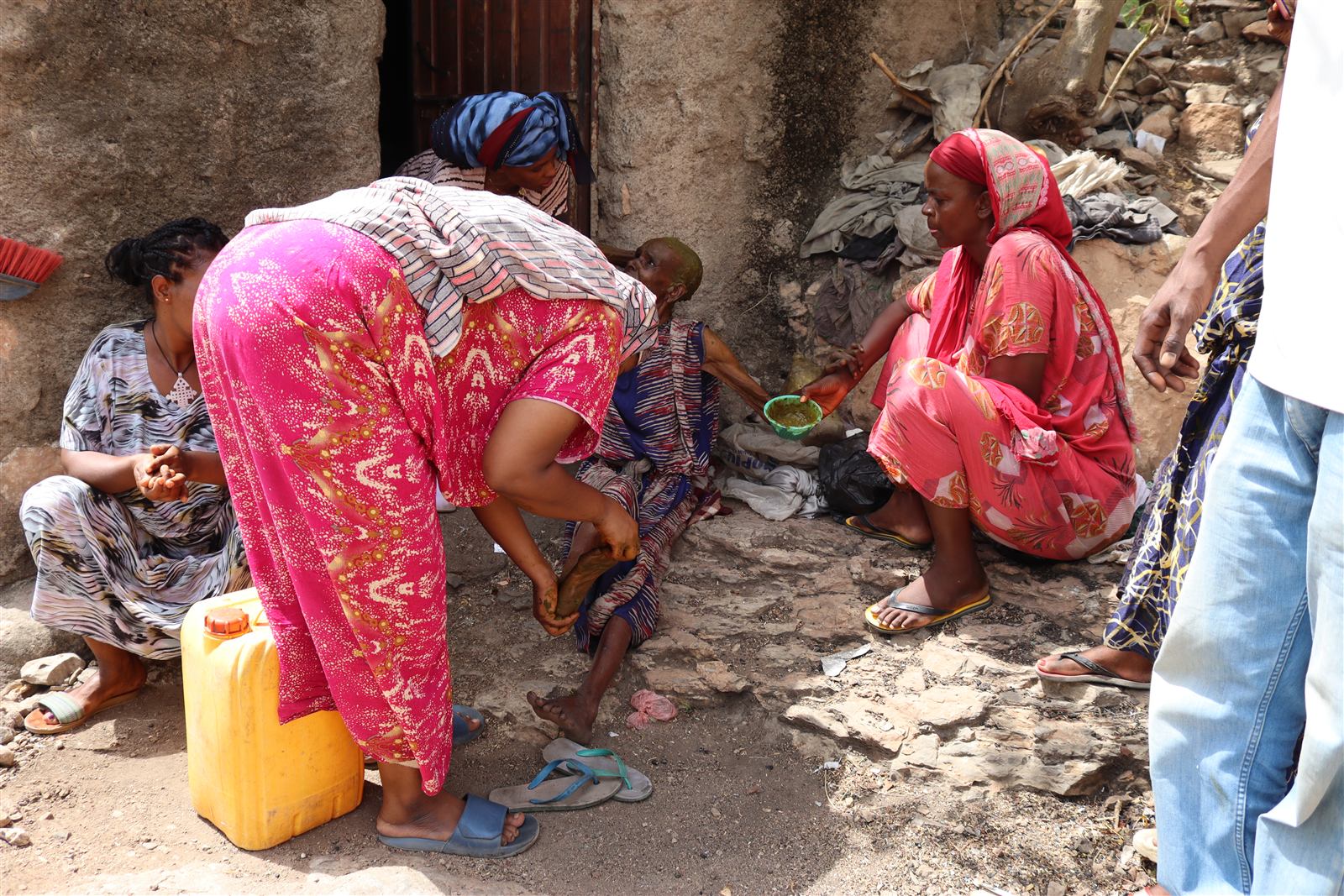 A woman kneels to tend to the elderly woman while another hands a bowl to a second seated woman