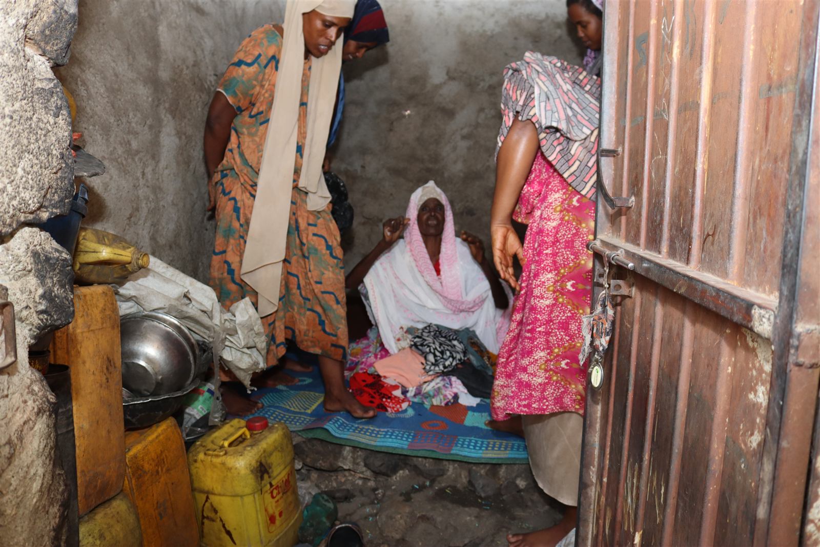 Inside a small room, two women attend to an elderly woman seated on a floor mat